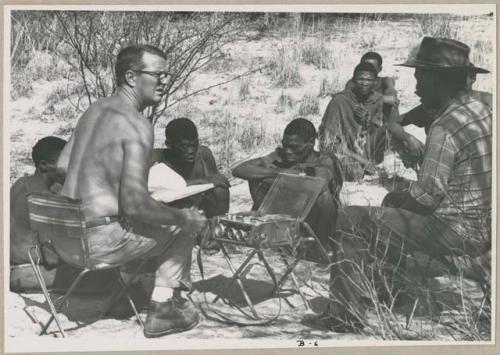 Wilhelm Camm and Nicholas England in chairs with a sound recorder interviewing a group of men sitting on the ground (print is a cropped image)