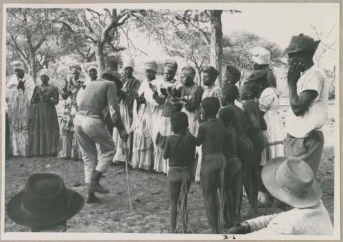 Group of women clapping, standing in a line with Judas Kangengi standing in the middle, preparing to sing; people watching (print is a cropped image)