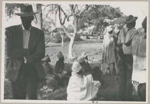 Man standing with his arms crossed; a group of women sitting and a group of people standing in the background (print is a cropped image)