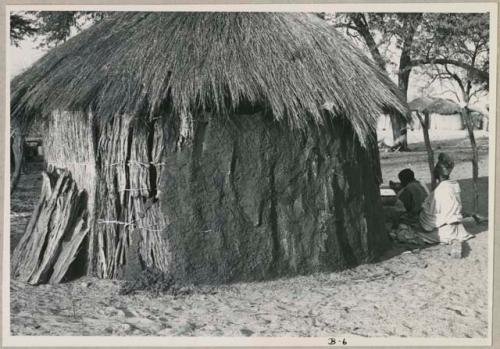 Woman and another person sitting beside a hut, seen from a distance (print is a cropped image)