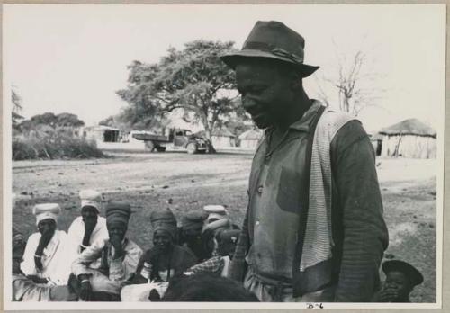Kangengi standing and listening to Nick England play back recordings; women sitting in the background (print is a cropped image)