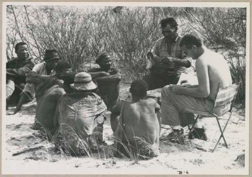 Wilhelm Camm and Nicholas England sitting in chairs with a sound recording machine taking notes, interviewing a group of men sitting on the ground (print is a cropped image)