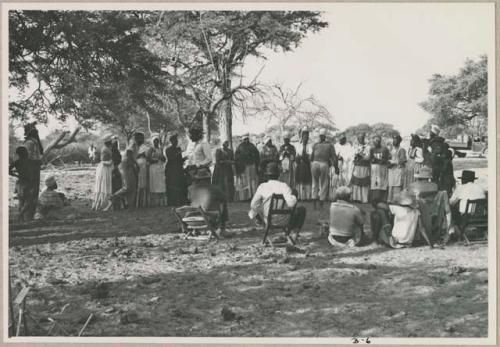 Group of women clapping, standing in a line with Judas Kangengi standing in the middle, preparing to sing; people watching (print is a cropped image)