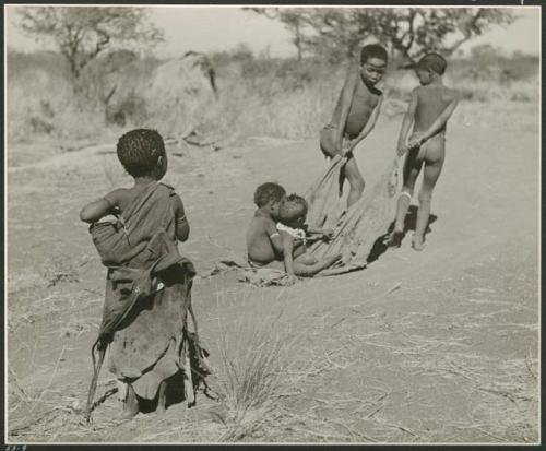 "Pulling on Kaross / Pulling on Gemsbok Horns / Carrying on Kaross "Airplane".": Tsamgao (≠Toma's son) and an unidentified boy pulling two babies on a kaross, with Be watching (print is a cropped image)