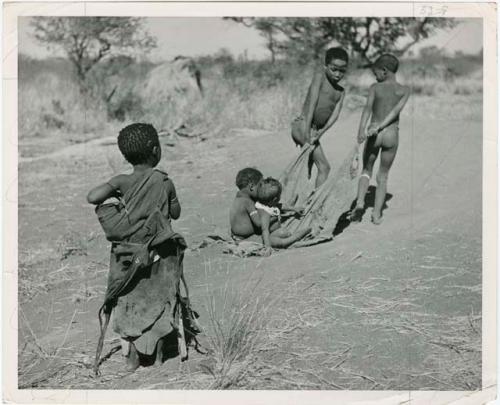 "Pulling on Kaross / Pulling on Gemsbok Horns / Carrying on Kaross "Airplane".": Tsamgao (≠Toma's son) and an unidentified boy pulling two babies on a kaross, with Be watching (print is a cropped image)