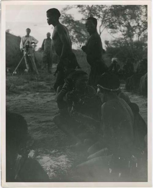 "Misc. prints with no numbers":  /Ti!kay and another man dance by groups of people sitting, with John Marshall and Laurence Marshall in the background (print is a cropped image)