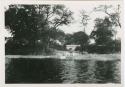 "1950 - 300 series duplicates": People standing along shore of Okavango River, with building in background