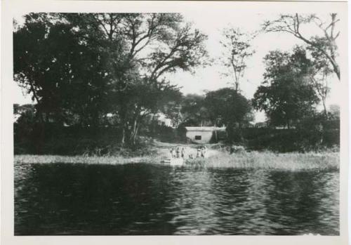 "1950 - 300 series duplicates": People standing along shore of Okavango River, with building in background