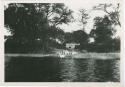 "1950 - 300 series duplicates": People standing along shore of Okavango River, with building in background