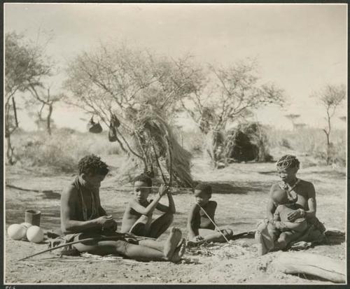 [No folder]: ≠Toma working on arrows and sitting with family members Tsamgao, /Gaishay and !U, holding Norna on her lap (print is a cropped image)