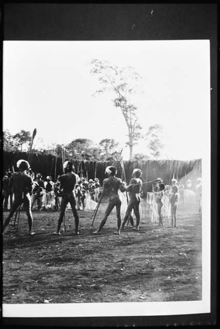 [No folder]: Group of people standing, including men wearing headdresses, view from behind, with a wall or fence in the background