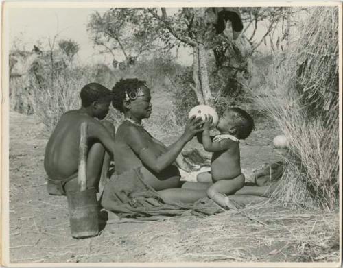 !Ungka (≠Toma's sister), with her daughter, Sa≠gai, sitting on her knees drinking from an ostrich egg shell; Tsamgao (Bau's husband) sitting beside her (print is a cropped image)