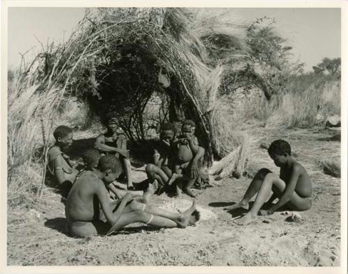 Gau of Band 2 sitting with his family in front of a skerm, including ≠Toma (his son with Be), his son !Naishi, Be (his first wife), Gau, Khuan//a (his second wife), /Qui (his son with Khuan//a), /Gunda (Khuan//a's son with her first husband), and ≠Gao (his son with Be) (print is a cropped image)