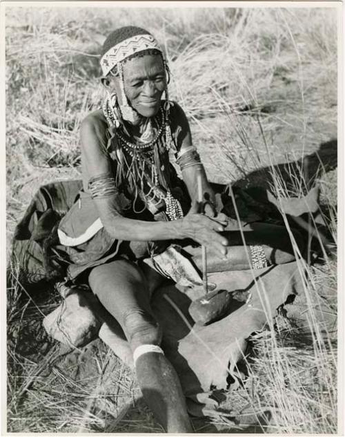 "Old /Gam" sitting and drilling holes in ostrich eggshell beads with her drill (print is a cropped image)