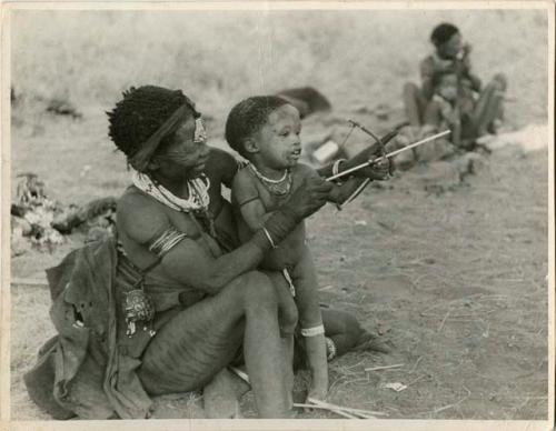 Di!ai showing her son, Debe, how to hold a toy bow and arrow (print is a cropped image)