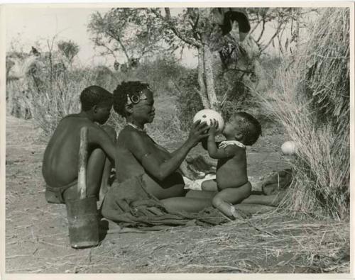 !Ungka (≠Toma's sister), with her daughter, Sa≠gai, sitting on her knees drinking from an ostrich egg shell; Tsamgao (Bau's husband) sitting beside her (print is a cropped image)