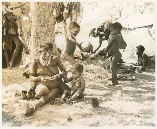 /Gaishay ("Gao Medicine's" and Di!ai's son), Bau ("Crooked /Qui's" and //Khuga's daughter) picking up the cooked wildebeest head; Di!ai and Debe sitting in the foreground and unidentified people in the background (print is a cropped image)