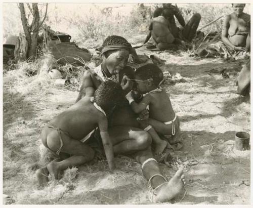 Di!ai sitting in a werft, stringing beads while nursing /Gaishay and Debe, and //Khuga nursing Bau in the background; unidentified people to the right (print is a cropped image)