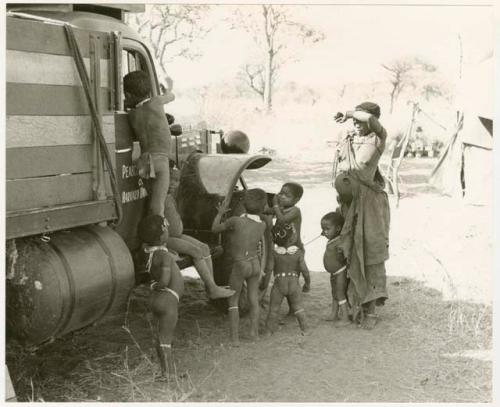 Children playing around the expedition truck while Di!ai stands beside them; the expedition camp is in the background (print is a cropped image)