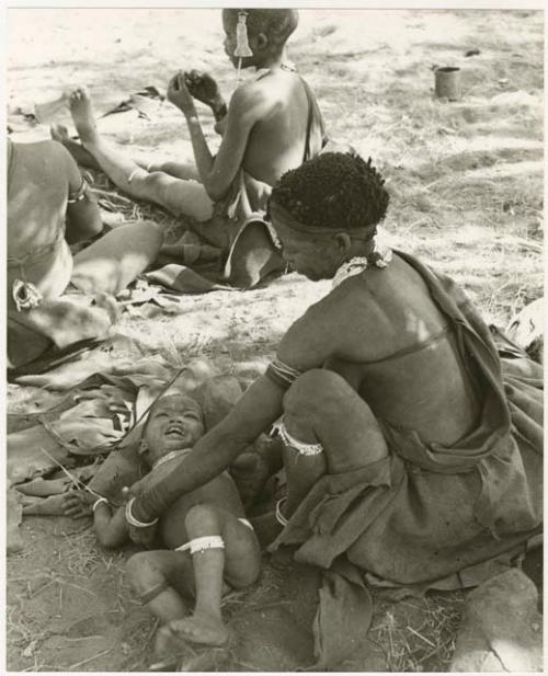 Di!ai sitting and reaching to pick up her son, Debe, with unidentified people in the background (print is a cropped image)