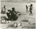 Khuan//a (Gau's wife) sitting with ostrich egg shells and children playing in the water behind her (print is a cropped image)