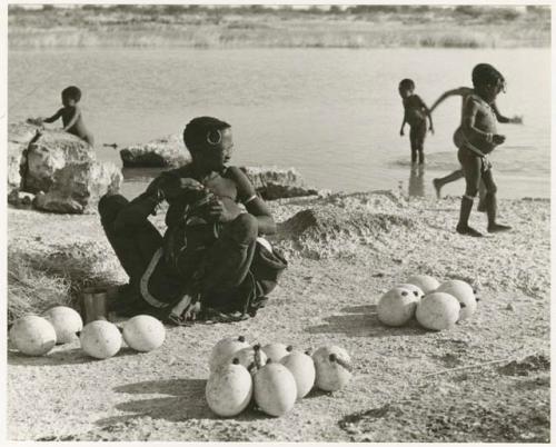 Khuan//a (Gau's wife) sitting with ostrich egg shells and children playing in the water behind her (print is a cropped image)