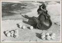 Khuan//a (Gau's wife) placing ostrich egg shells wrapped in grass in her kaross and her child standing near her (print is a cropped image)