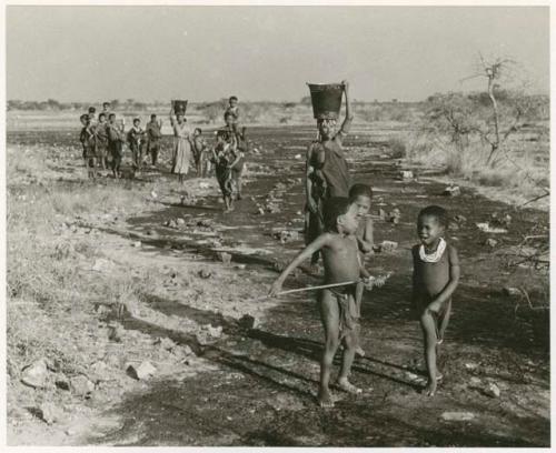 Women and children returning from Nama Pan with water; Di!ai and !Ungka carrying buckets on their heads, distant view (print is a cropped image)
