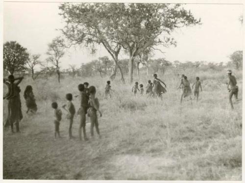 Girls playing N!owa t/ama (melon tossing game) / tamah n!o’an (ball game); other children are watching and running, distant view (print is a cropped image)