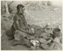 Woman pounding bone and red powder in her kaross with a stone (print is a cropped image)