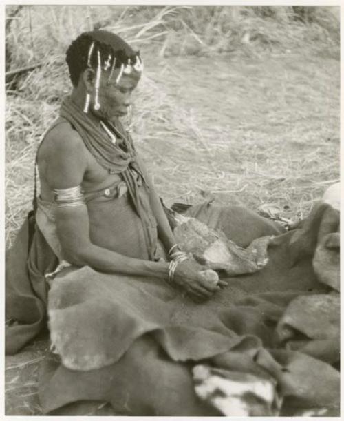 Woman pounding bone and red powder in her kaross with a stone (print is a cropped image)