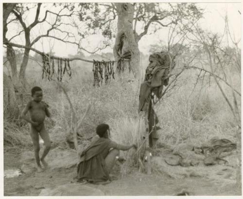 Woman and boy in their living space; biltong hanging from a wooden stick above them (print is a cropped image)