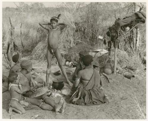 Group sitting in a werft by a fire; left to right: "Old /Gasa," Dam ("Old /Gasa's" son-in-law; ≠Gisa's husband) playing a //guashi, an unidentified girl kneeling next to him, a woman, mostly likely /Goishay, and ≠Gao ("Old /Gasa's" son; Gau's brother) (print is a cropped image)