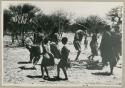 [No folder title]: People performing the Eland Dance, with Twi imitating the sideways motion of an eland's hind legs and playing up to ǂKxoba (from color slide 2001.29.8658)