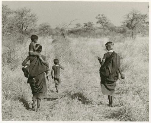 Di!ai walking from the back with Debe sitting on her shoulder while /Gaishay runs ahead; another unidentified woman is walking beside her (print is a cropped image)