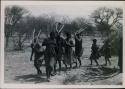 [No folder title]: Group of people performing the Eland Dance; !Ani leading a line of people, men holding wooden horns to their heads
