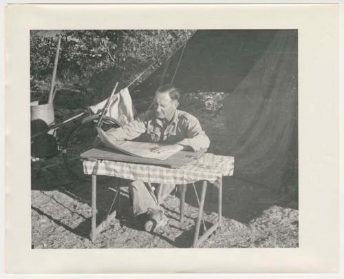 "1950's 400 series": Man working at a table in front of a tent (print is a cropped image)