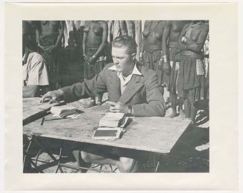 "1950 '400 Series' / 87 B/W prints (some duplicates) / Ovamboland": Group of people standing behind a man sitting at a table (print is a cropped image)