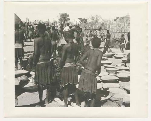 "1950 '400 Series' / 87 B/W prints (some duplicates) / Ovamboland": Group of people sitting and standing next to baskets of grain on ground  (print is a cropped image)