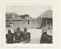 "1950 '400 Series' / 87 B/W prints (some duplicates) / Ovamboland": People sitting inside kraal, with huts in background (print is a cropped image)