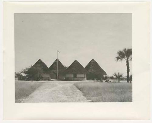 "1950 '400 Series' / 87 B/W prints (some duplicates) / Ovamboland": Group of people standing and sitting next to four round buildings with pointed roofs and flag (print is a cropped image)