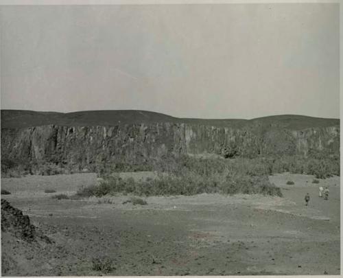 "1950 400 series  40 prints / Kaokoveld": Three men walking, with cliff in background (print is a cropped image)
