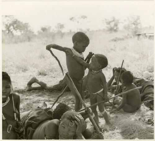 [No folder title]: "The visiting girl" sitting, !U lying down with Norna against her, Di!ai's sons /Gaishay and Debe playing with sticks, and Bau sitting with /Naoka (Di!ai's co-wife) (print is a cropped image)