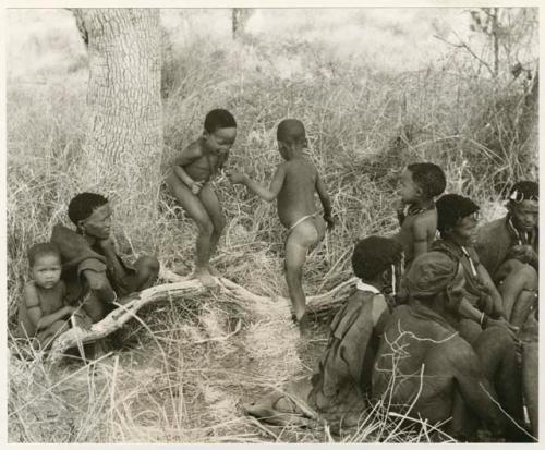 [No folder title]: "Little ≠Gao" and an unidentified boy playing on a log amid a seated group of people (print is a cropped image)