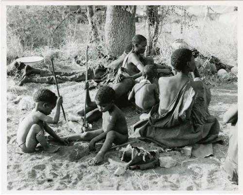 [No folder title]: !Ungka Norna and Bau playing with digging sticks next to a group of people sitting (print is a cropped image)