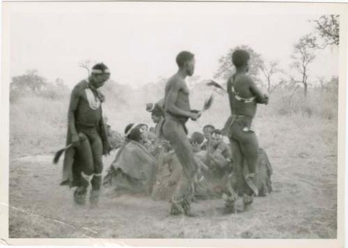 [No folder title]: Three men, including /Gunda and ≠Gao, dancing around a group of seated women singing; medicine man leaning over "Gao Medicine" far back behind (print is a cropped image)