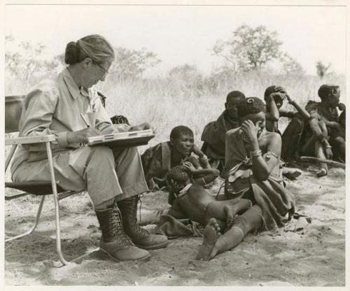 [No folder title]: Lorna Marshall taking notes, !U, !Ungka Norna, /Naoka ("Gao Medicine's" first wife), and other unidentified people in the background (print is a cropped image)