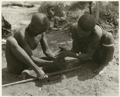 [No folder title]: Dam and ≠Gao creating a fire with firesticks; unidentified people in the background (print is a cropped image)