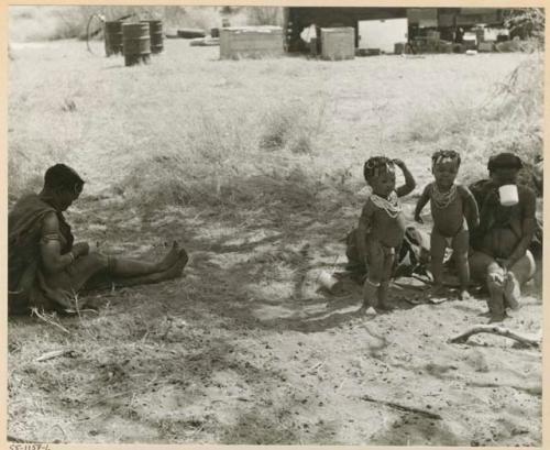 [No folder title]: Khuan//a sitting to the left, her youngest son ≠Toma standing with an unidentified girl, and two unidentified women with the expedition camp in the background (print is a cropped image)