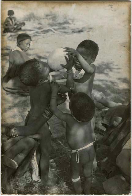 [No folder title]: Debe holding Bau's arms as she drinks from an ostrich eggshell held by /Gaishay while an unidentified, young woman steadies them; unidentified women sit in the background (print is a cropped image)